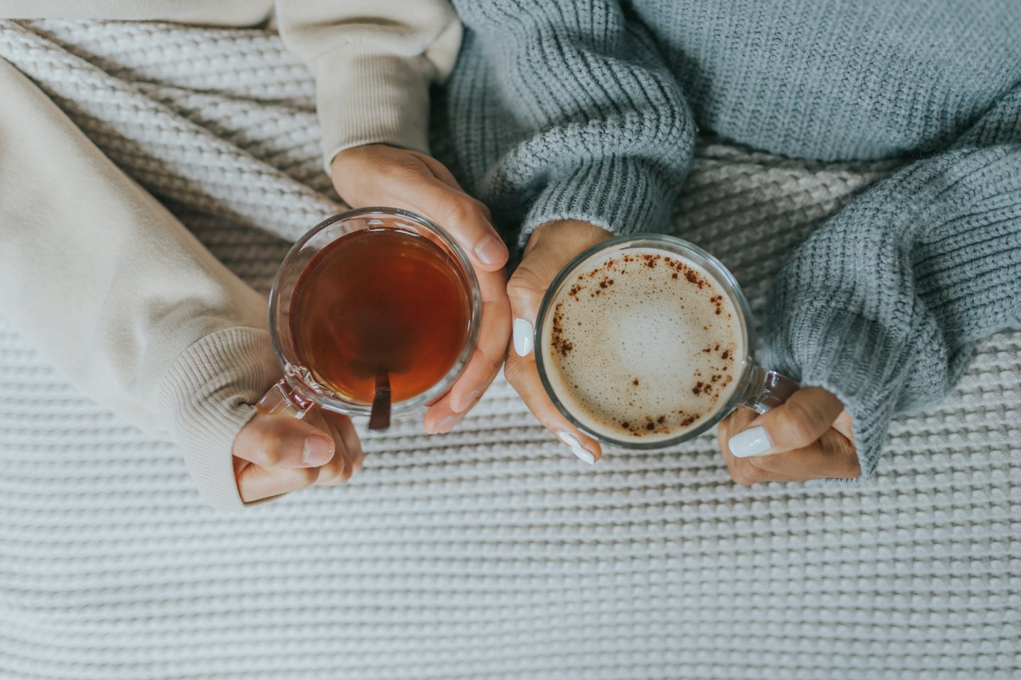Two people hold mugs of hot drinks on a knitted blanket.