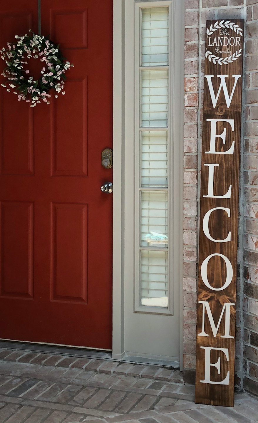 A floral wreath hangs on a red door by a wooden welcome sign.